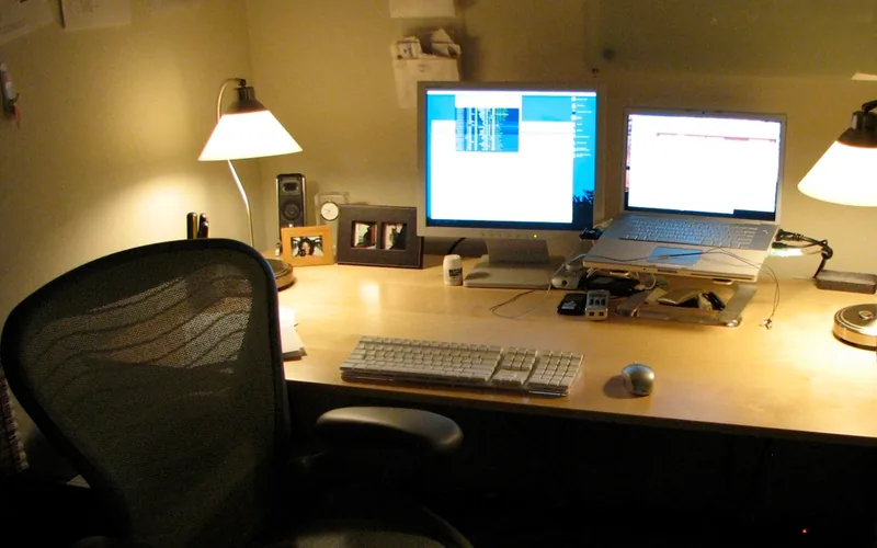 Mesa de trabajo de noche con una lámpara de tulipa blanca encendida a cada lado de un monitor y un portátil; delante, una silla de malla