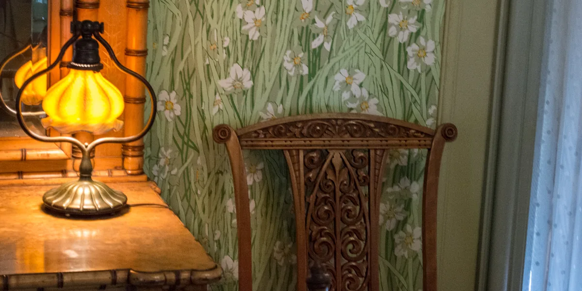 Lámpara de sobremesa con tulipa de vidrio ámbar encendida sobre un mueble de madera con espejo, junto a una silla tallada y una ventana con visillo